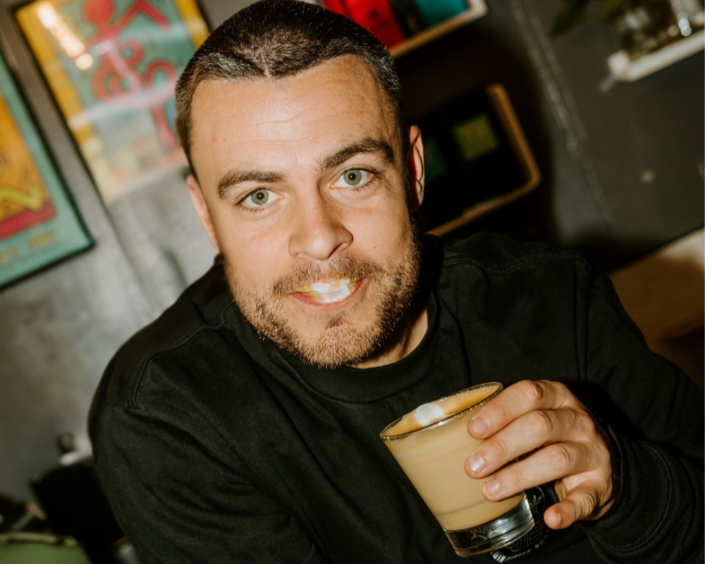 Man from The Bean Cartel holding a coffee cup in a casual setting with colorful artwork in the background